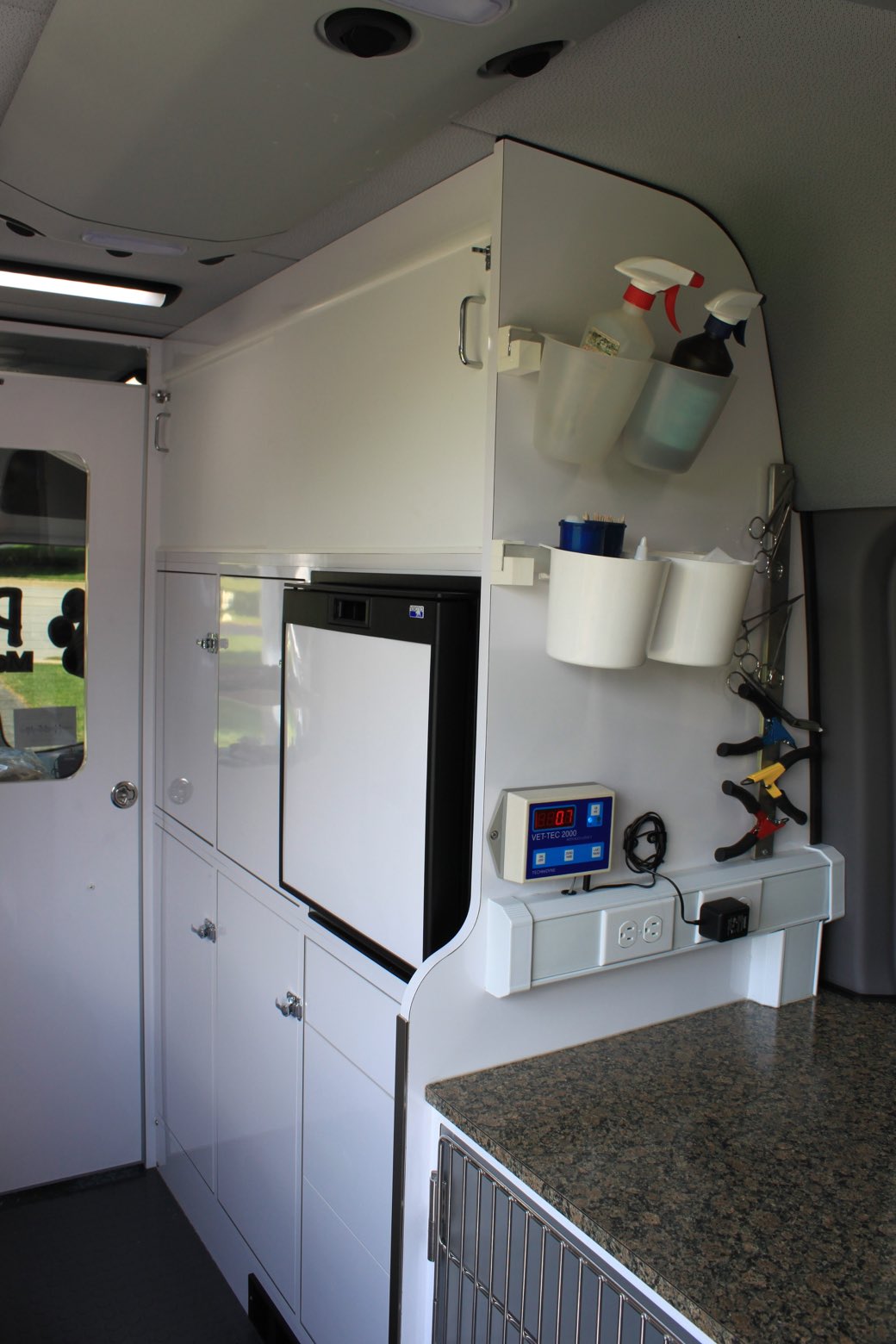 The fitted interior of a mobile veterinary van: cabinets, a refrigerator, a dental unit and a stainless kennel under the counter